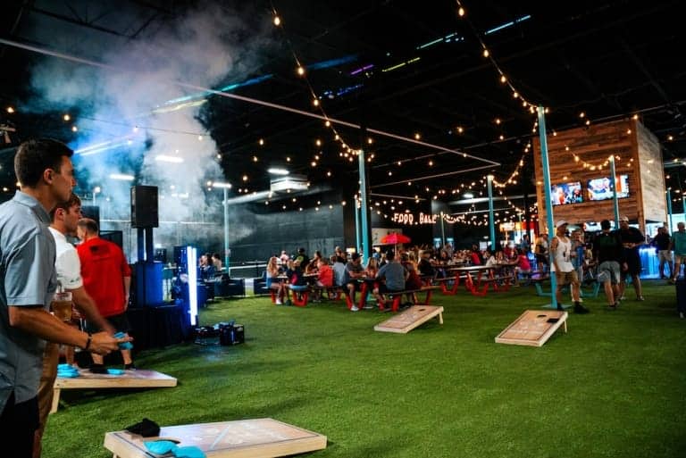 Group of people posing for a photo, cornhole game in background - Pickle-Pins